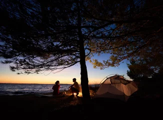 Fotobehang Romantic supper on lake shore at night. Tourist tent under trees and young family, man and woman at campfire on dark blue evening sky and clear water background. Tourism and camping concept. © anatoliy_gleb