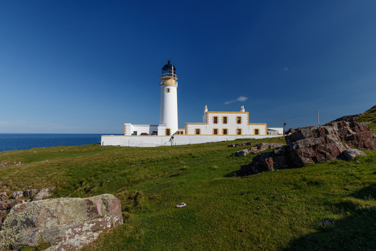 Rua Reidh Lighthouse with keepers house and walls on grassy hillside along the coast. Rocks in the foreground and blue sky.