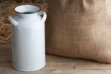 Mockup of a white vintage milk jug on a wooden table next to straw and a burlap sack.