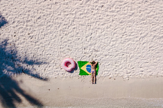 Aerial view of a young woman with a brazilian flag on the beach. Crystal clear water, tropical vacation scenario, coconut tree and boats. Drone view