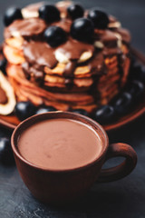 Hot chocolate drink in a cup and pancakes with banana, chocolate sauce and grapes in plate, on dark background