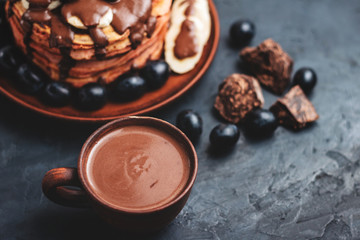 Hot chocolate drink in a cup and pancakes with banana, chocolate sauce and grapes in plate, on dark background