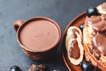Hot chocolate drink in a cup and pancakes with banana, chocolate sauce and grapes in plate, on dark background