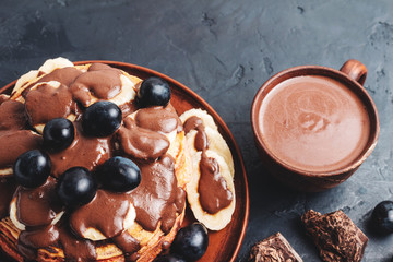 Pancakes with banana, chocolate sauce and grapes in a plate and hot chocolate drink in a cup on dark background