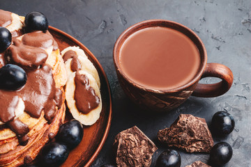 Hot chocolate drink in a cup and pancakes with banana, chocolate sauce and grapes in plate, on dark background