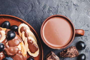 Hot chocolate drink in a cup and pancakes with banana, chocolate sauce and grapes in plate, on dark background, top view