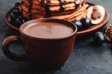 Hot chocolate drink in a cup and pancakes with banana, chocolate sauce and grapes in plate, on dark background