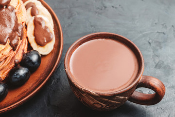 Hot chocolate drink in a cup and pancakes with banana, chocolate sauce and grapes in plate, on dark background
