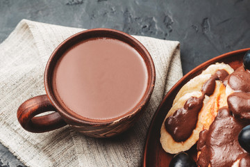 Hot chocolate drink in a cup and pancakes with banana, chocolate sauce and grapes in plate, on dark background