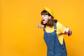 Angry irritated girl teenager in french beret, denim sundress looking aside clenching fists screaming isolated on yellow wall background. People sincere emotions lifestyle concept. Mock up copy space.