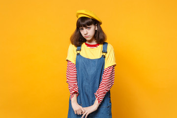 Portrait of offended disconcerted girl teenager in french beret, denim sundress standing isolated on yellow wall background in studio. People sincere emotions, lifestyle concept. Mock up copy space.