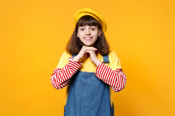 Portrait of smiling girl teenager in french beret, denim sundress holding hands near face isolated on yellow wall background in studio. People sincere emotions, lifestyle concept. Mock up copy space.