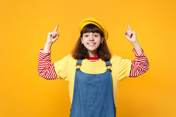 Portrait of smiling girl teenager in french beret denim sundress pointing index fingers up isolated on yellow wall background in studio. People sincere emotions, lifestyle concept. Mock up copy space.