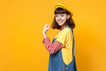 Portrait of beautiful girl teenager in french beret, denim sundress showing biceps, muscles isolated on yellow wall background in studio. People sincere emotions lifestyle concept. Mock up copy space.