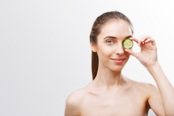 Portrait of brunette woman covering eye with cucumber slice isolated on white background