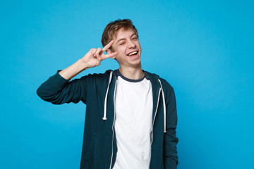 Portrait of cheerful funny young man in casual clothes blinking and showing victory sign isolated on blue wall background in studio. People sincere emotions, lifestyle concept. Mock up copy space.