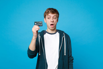 Shocked young man in casual clothes holding, looking on credit bank card, keeping mouth wide open isolated on blue background in studio. People sincere emotions, lifestyle concept. Mock up copy space.
