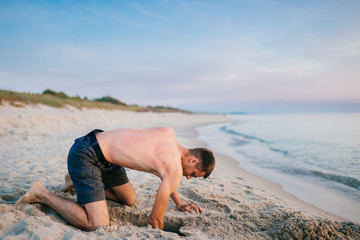 Young topless barefoot man in shorts digging hole by hands on beach beyond sea in summer evening. Funny boy playing on beach with sand on desolate cost near ocean ike child. Vacation leisure and hobby