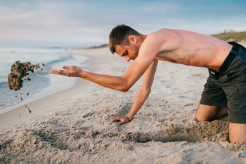 Young topless barefoot man in shorts digging hole by hands on beach beyond sea in summer evening. Funny boy playing on beach with sand on desolate cost near ocean ike child. Vacation leisure and hobby