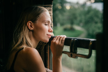 Mood atmospheric lifestyle portrait of young beautiful blonde hair girl looking out of window from riding train. Pretty teen enjoying beauty of nature from moving train car in summer. Travel concept