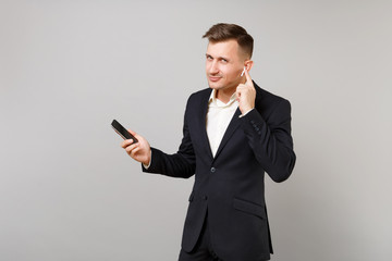 Successful young business man in classic suit with wireless earphones listening music, holding mobile phone isolated on grey background. Achievement career wealth business concept. Mock up copy space.