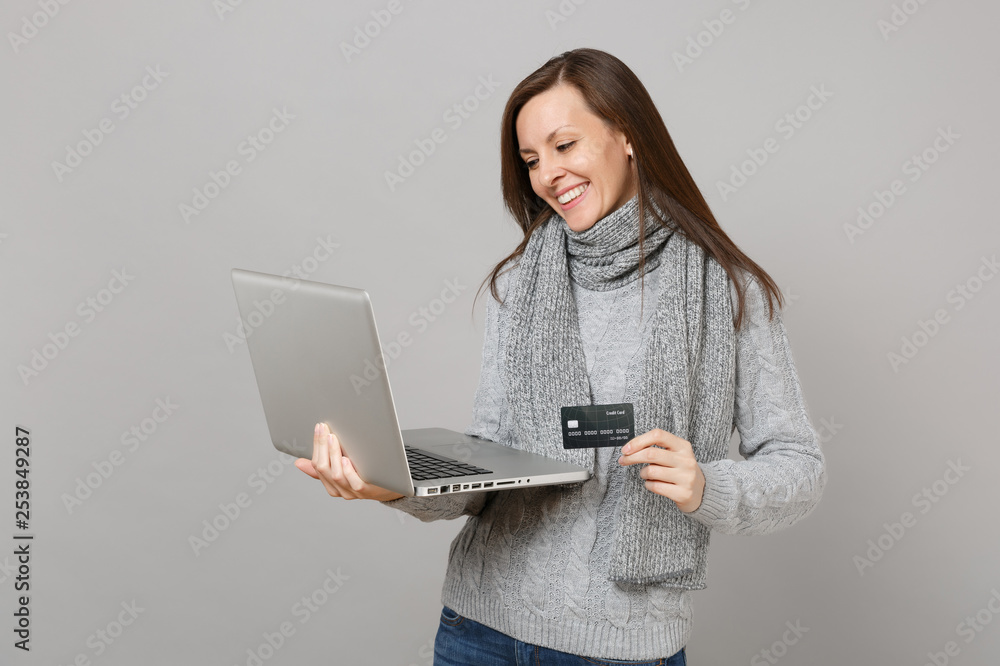 Wall mural Smiling young woman in gray sweater, scarf working on laptop pc computer holding credit bank card isolated on grey wall background. Healthy lifestyle, online treatment consulting, cold season concept.