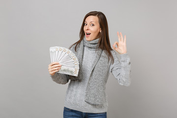 Amazed young woman in gray sweater scarf showing OK gesture hold lots bunch of dollars banknotes cash money isolated on grey background. Healthy fashion lifestyle people emotions, cold season concept.