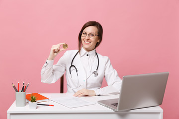 Female doctor sits at desk works on computer with medical document hold coin in hospital isolated on pastel pink wall background. Woman in medical gown glasses stethoscope. Healthcare medicine concept