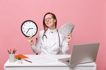 Female doctor sit at desk work on computer with medical document hold money in hospital isolated on pastel pink wall background. Woman in medical gown glasses stethoscope. Healthcare medicine concept.