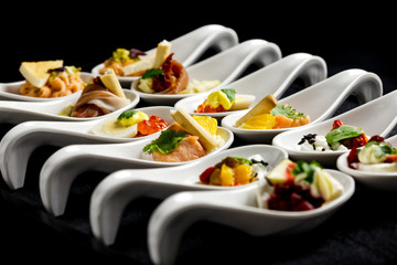 Two rows of canapes on white ceramic spoons. Black background.