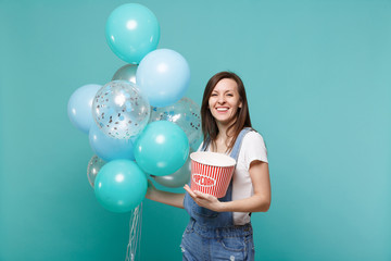 Smiling joyful young woman in denim clothes holding bucket of popcorn, celebrating with colorful...