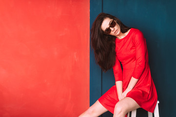 Beautiful fashion girl with long hair, spanish appearance in sunglasses and red elegant dress posing on blue red wall in studio. Amazing babe in suglasses with black hair. Hairstyle advertsing.
