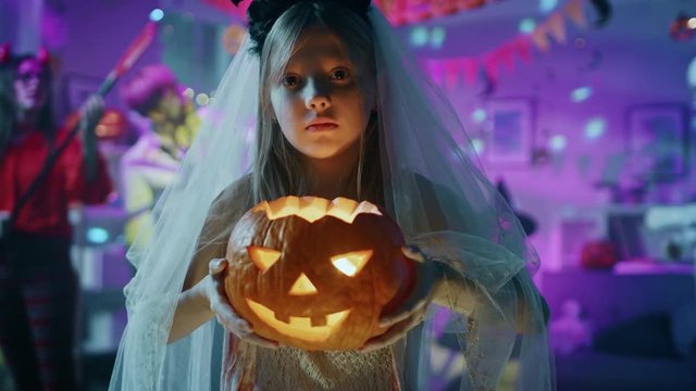 Halloween Costume Party: Smiling Little Girl in a Bloody White Bride Dress Holding Burning Pumpkin Head. Zombie, Blood Thirsty Dracula, Mummy, Bewitching Witch and Dazzling She Devil Dance