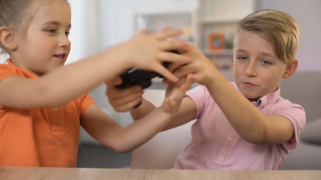 Little girl disturbing brother playing console game, children fighting joystick