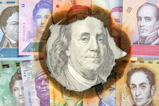 Concept of hyperinflation Venezuela. Burned Venezuelan money with the image of statesmen, through hole in which you can see an unburned one hundred dollar bill with Franklin. Close up.