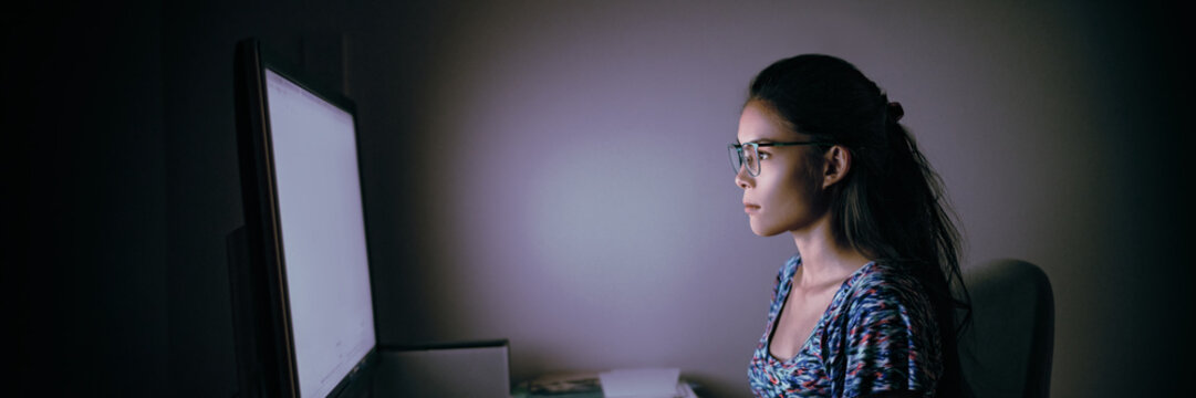 Computer online internet addiction young woman staring at computer screen late at night in dark room. Asian businesswoman working overtime at office desk using blue light glasses eye strain care.