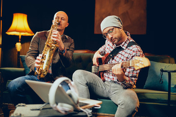 Two talented musicians playing bass gutar and sax while sitting on couch in home studio.
