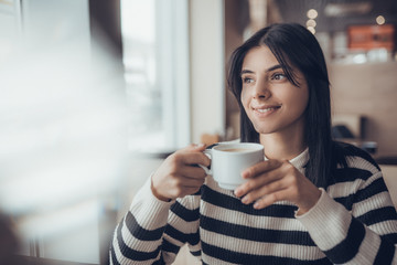 Beautiful caucasian girl in casual clothes with coffe in hands smiling while sitting in cafe. success.