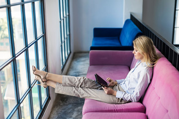 Blond businesswoman sitting on violet couch at the window using tablet
