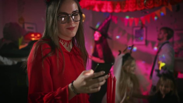 Halloween Costume Party: Seductive She Devil with Trident Uses Smartphone. In the Background Group of Monsters Having Fun, Dancing Under Disco Ball.