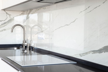 Close up of white glossy kitchen with black quartz countertop and marble tile backsplash. Build-in hidden incorporated hood and undermounted sink.  - obrazy, fototapety, plakaty