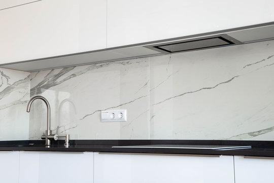 White glossy kitchen with black quartz countertop and marble tile backsplash. Build-in hidden incorporated hood and undermounted sink. 