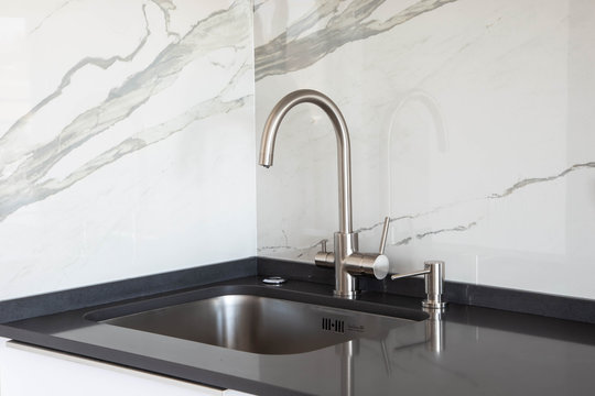 Close up of a undermounted sink and mixer in white and black kitchen design with marble tile backsplash and dark grey quartz countertop. 