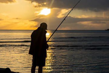 An unrecognizable man fishes while a beautiful sunset creates a stunning silhouette.