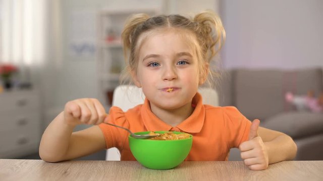 Hungry female kid eating cornflakes with milk and showing thumbs up, breakfast