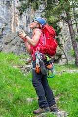 Happy climber girl with a red backpack, quickdraws, climbing shoes, chalk bag attached to her harness, making a wish for succeeding on a sport climbing route, while looking up at the stone wall.