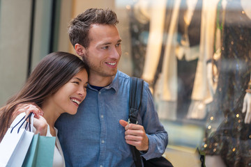 Multiracial couple shopping together looking at clothing in store window walking in city street. Happy asian woman holding shopping bags with caucasian man.