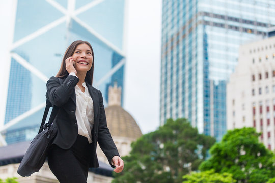 Asian Businesswoman Talking On Mobile Phone Walking In Hong Kong City Street To Office, Skyscrapers Cityscape Background. Young Woman On Smartphone Happy. Multiracial Chinese Caucasian Lady.