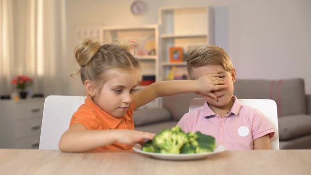 Little sister feeding brother with broccoli covering his eyes, joking friend