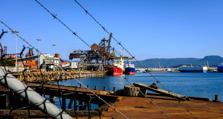 Ships loading coal and iron ore at industrial port taken through razor wire fence in foreground at...